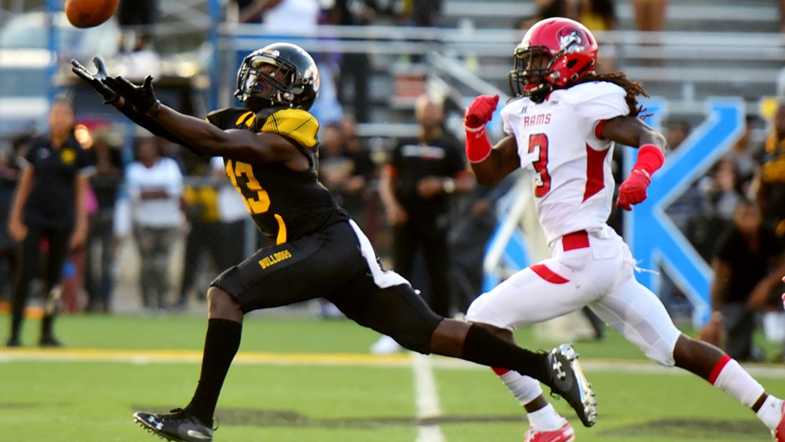 Receiver runs to catch the footbal with his arms outstretched and an opposing player right behind him