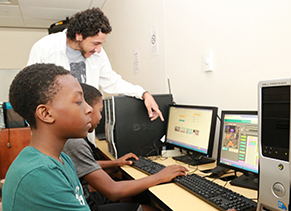 A boy uses a computer while a college student instructs another one